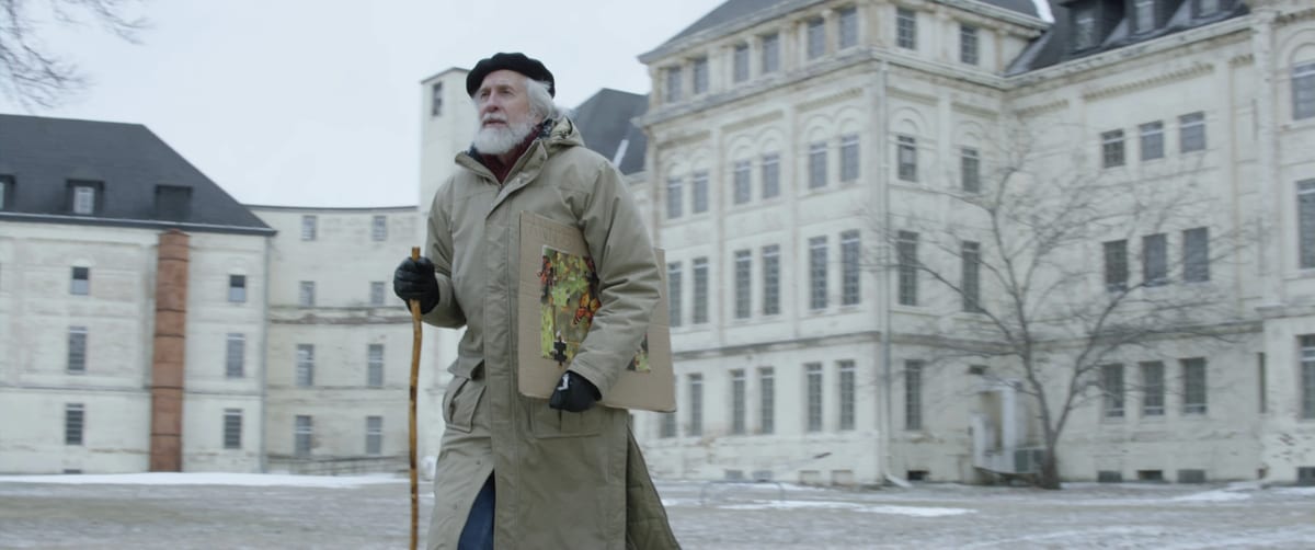 Cris Krusen in a long coat and beret, standing in the snow in front of a large stone hospital.