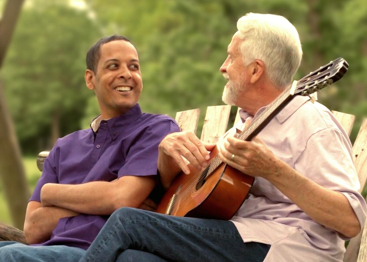 Two men on a garden bench, one playing an acoustic guitar.