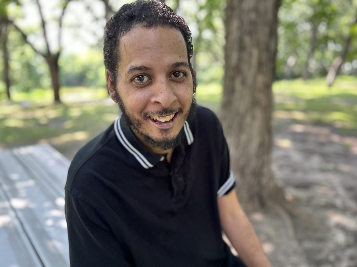 A man smiling outdoors under the trees.