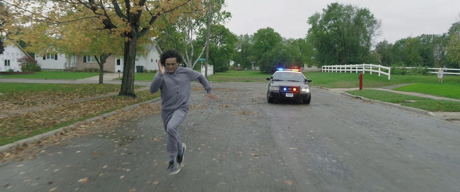 A young man running down a residential street, a police car behind him.