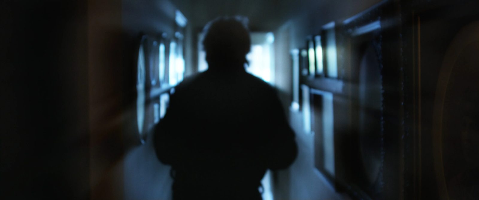 A silhouette walking down a dark hospital corridor toward a lit doorway.